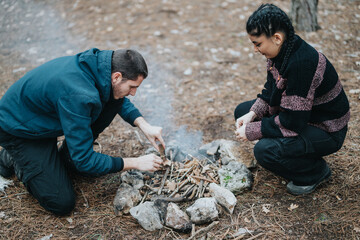 A man and a woman are kneeling by the campfire, arranging the wood for ignition during an outdoor...