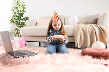Birthday celebration. Little girl blowing candle on piece of cake while having video call via...