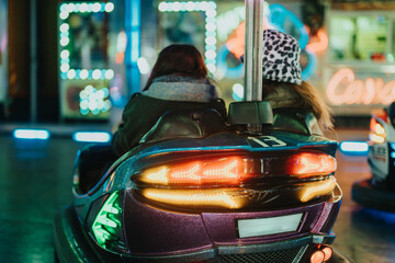 Two friends ride together in a bumper car during a festive evening at a colorful fairground.