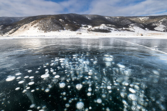 View of frozen methane bubbles trapped beneath the clear, icy surface, with distant, snow-dusted mountains under a bright sky, Baikal, Irkutsk Oblast, Russia.