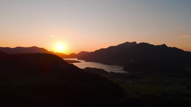 Slow-rising drone shot at golden hour unveils Lake Wolfgangsee and the village of Strobl, framed by the Salzburg Alps as the sun sets and pastel light reflects on tranquil water.