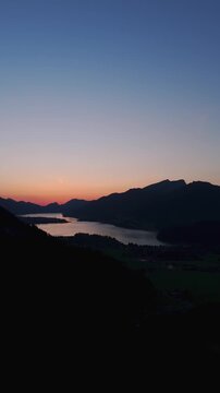 Vertical forward-moving drone glides over Lake Wolfgangsee toward Strobl town as soft post-sunset afterglow paints the Salzburg Alps in gentle orange-pink twilight.
