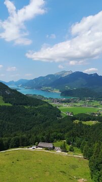 Vertical left-orbit drone shot sweeps above Lake Wolfgangsee, circling the alpine resort town of Strobl. Summer sunlight highlights turquoise water, green meadows and the surrounding Salzburg Alps.