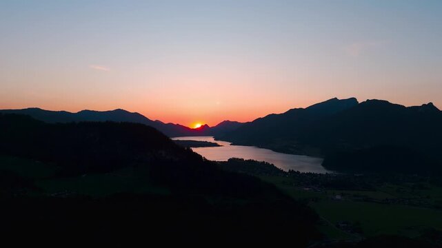 Right-orbit aerial circles Lake Wolfgangsee at dusk, framing Strobl village and the Salzburg Alps as gentle afterglow paints the horizon in pastel orange-pink and reflects on calm water.