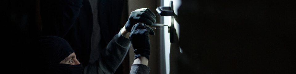 Burglar wearing black mask and gloves picking door lock with metal tool in dark environment,...