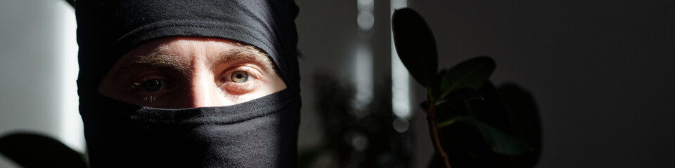 Portrait of male burglar wearing black balaclava looking into camera with serious expression,...