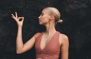 Close-up of calm woman making mudra hand gesture, eyes closed, standing against black rock,...