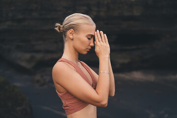 Close-up of woman pressing palms together in prayer mudra, standing still on dark sand, expressing...