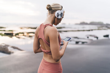 Blonde woman with headphones stands turned away, reading smartphone, surrounded by ocean and black...