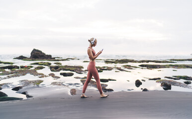 Full-body shot of woman walking barefoot in headphones and activewear across volcanic beach,...