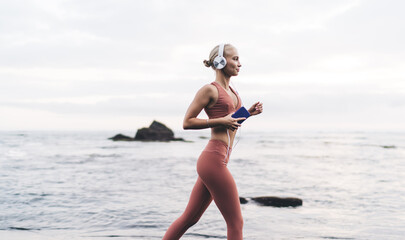 Blonde runner strides confidently with smartphone and headphones, barefoot along ocean, expressing...