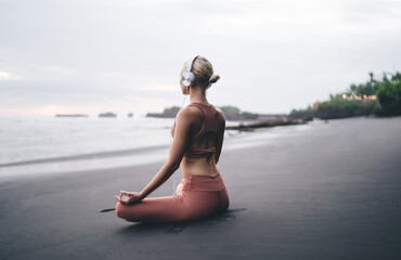 Woman in activewear sits at the edge of black beach, meditating in headphones, calmly looking...