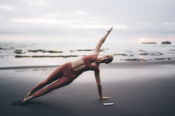 Blonde woman performs side plank yoga pose barefoot on volcanic beach during sunset, smartphone...