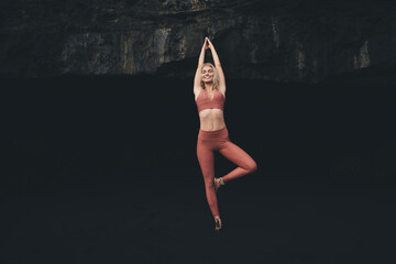 Young woman stands in tree pose, hands raised above head, smiling calmly in front of black rocky...