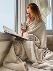 Ginger-haired woman, 30s, sitting on bed by the window in cozy covered in blanket, reading a book...