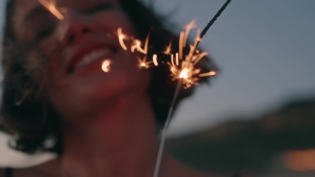 Closeup of young woman smiling and holding a sparkler at sunset. Sparkler burning brightly in the foreground. Happy, carefree, and joyful.