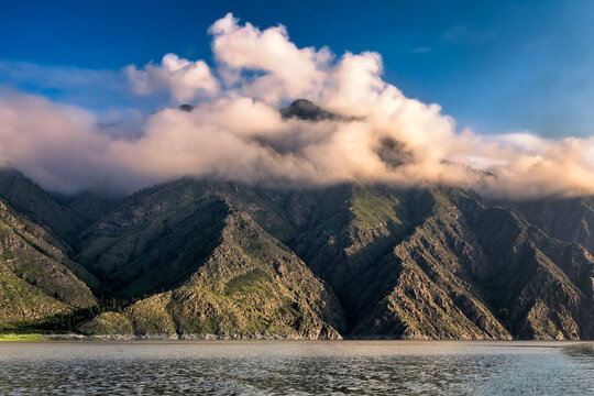 View of rugged, sun-kissed mountain peaks cloaked in ethereal, swirling clouds, reflected in the calm waters below, Sayan Mountain, Russia.
