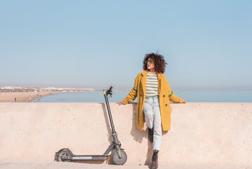 Woman in yellow coat with electric scooter enjoying sunny day at beach,sustainable urban mobility