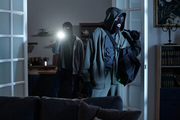 Two masked men entering dark room, one carrying large bag and other shining flashlight, both...