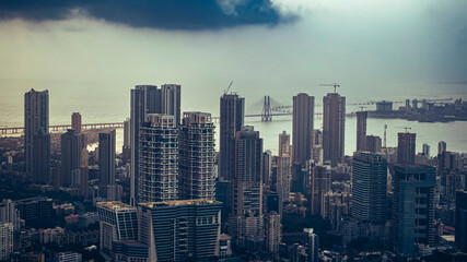 Aerial view of Mumbai city during a monsoon evening with dramatic, cloudy skies. Bandra-Worli Sea Link is visible behind high-rise buildings, a scenic urban and weather contrast.