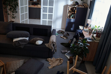 Living room scene showing scattered books, pillows, and clothing on floor near sofa and window,...