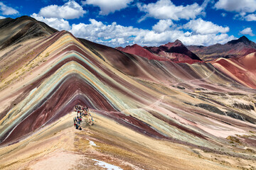 Vinicunca, Peru - 17 July 2025: Aerial view of the vibrant Rainbow Mountain, its striated slopes a tapestry of ochre, rust, and gold under a sky streaked with clouds.