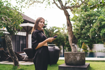 Confident young woman holds smartphone while standing in resort garden, dressed in black, enjoying...