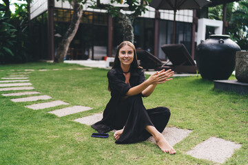 Barefoot woman clapping hands joyfully on grass with smartphone nearby—representing happiness,...