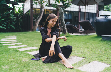 Woman lowers gaze with soft smile, sitting cross-legged barefoot in lush garden—symbol of...