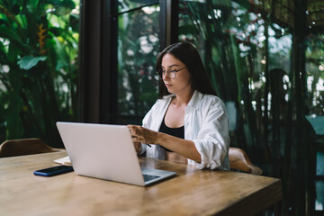 Focused young woman types on laptop indoors, surrounded by greenery, symbolizing digital...