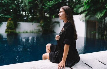 Woman in black sitting at pool edge, looking away thoughtfully—embodying quiet power, focus and...