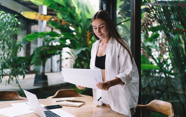 Freelancer woman works remotely in tropical space, reviewing printed reports while immersed in lush...