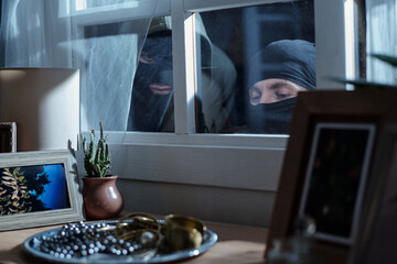 Two masked men peering through window, observing valuables on table inside house, faces partially...