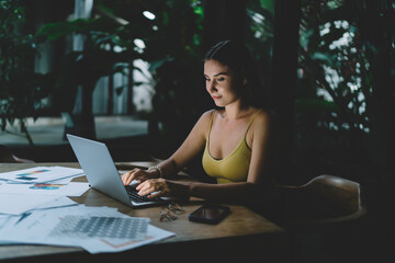 Young female freelancer typing on laptop, focused on screen in dark tropical interior, working...