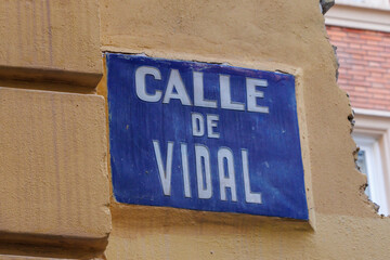 Street sign for Calle de Vidal, displaying vibrant blue and white colors, set against a yellow wall...
