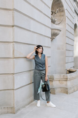 Trendy woman in gray vest and pants posing near stone wall with black handbag.