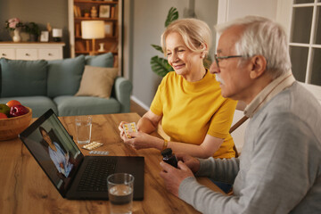 An elderly couple sits at their wooden table, showing their medications to a Doctor on their laptop screen. They are in their living room during the daytime.