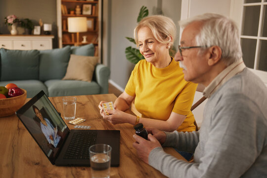 An elderly couple sits at their wooden table, showing their medications to a Doctor on their laptop screen. They are in their living room during the daytime. - Powered by Adobe