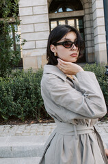 Elegant woman posing confidently in trench coat near historical building with greenery background