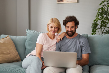 A couple sits closely on a comfortable couch, smiling as they share a laptop. Their relaxed...