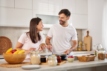 A couple is joyfully interacting while cooking in a modern kitchen. They are sharing smiles and laughter, preparing a healthy breakfast surrounded by fresh ingredients.