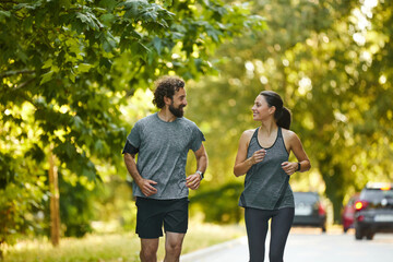 Two individuals are jogging side by side along a tree-lined pathway, smiling and engaging in...