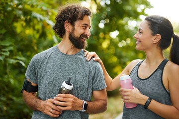 In a vibrant park, a man and woman share a joyful moment post-jogging. They hold water bottles and...