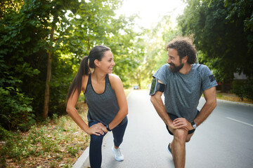 Two individuals perform warm-up stretches side by side on a serene road. Lush trees provide shade...