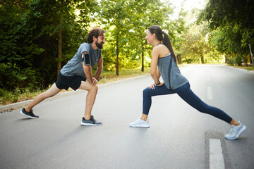 Two people engage in stretching exercises on a serene road lined with trees in the morning. Their...