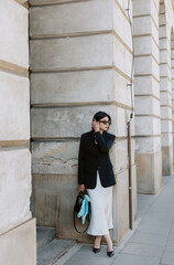 Woman leaning on stone wall in black blazer and white skirt with relaxed elegance