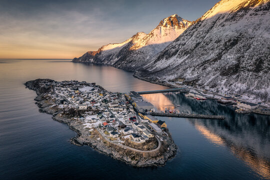 Aerial view of husoy village nestled on a rocky outcrop, kissed by the golden glow of sunrise against snow-dusted mountains and serene waters, Norway.