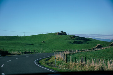 Scenic roadside view in rural New Zealand with rolling green hills, wire fencing, and a curving...