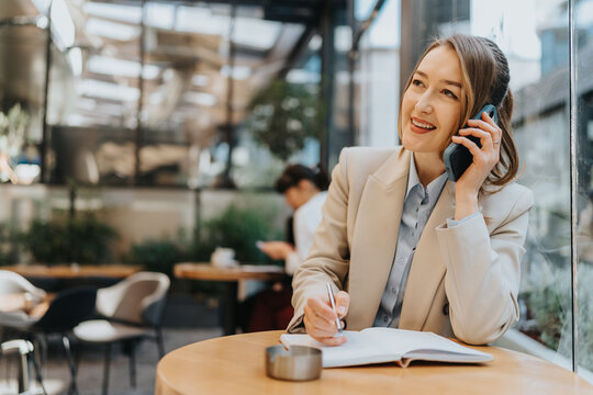 A businesswoman in a well-lit modern cafe jotting down ideas while talking on the phone, showcasing multitasking and professional communication.