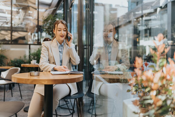 A businesswoman holding a phone and writing in a notebook, working in a modern outdoor cafe during sunlight hours.